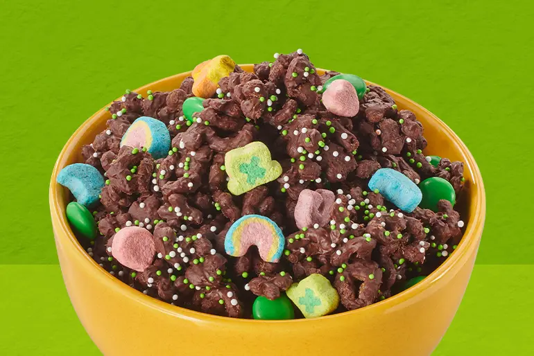 Un bol amarillo con mix de snacks de chocolate de Lucky Charms™ en un fondo verde.