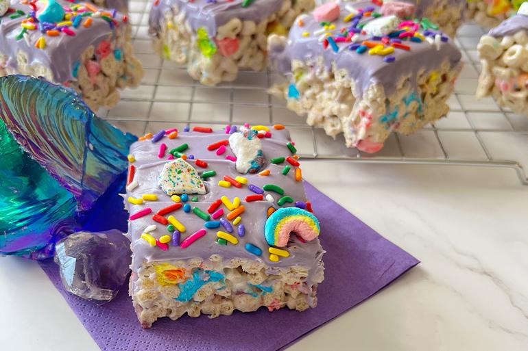 Receta de cereal de gemas mágicas congeladas de Lucky Charms™ servida en una servilleta morada posicionada en frente de otras barras de cereal en una bandeja de hornear.