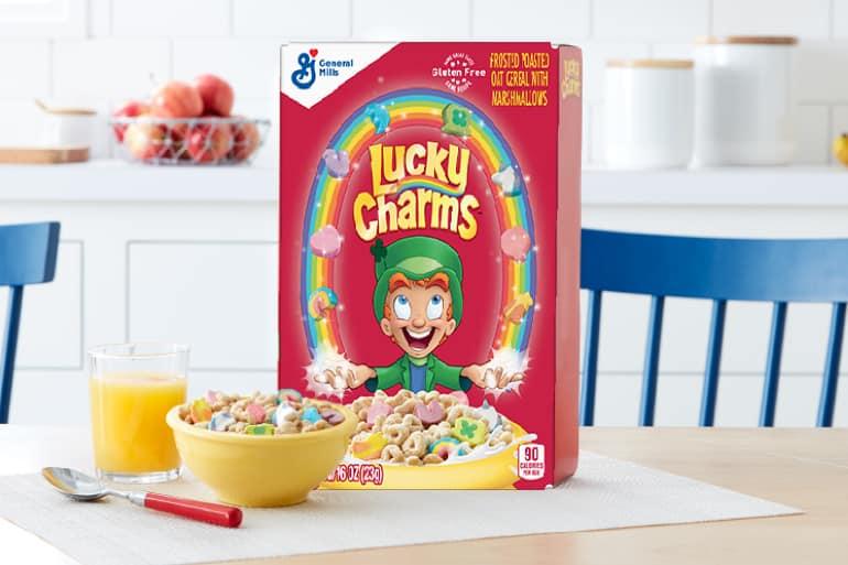Caja de cereal de avena tostada de Lucky Charms™ frosted con marshmallows en una mesa con bebidas y una cuchara.