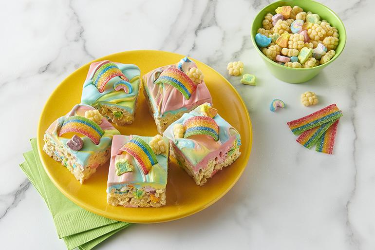 Cuadrados de barras de cereal glaseadas en un plazo amarillo con Lucky Charms™ Marshmallow Clusters en el fondo.