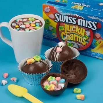 Cuatro bolas de chocolate con Lucky Charms y una caja de Swiss Miss® Lucky Charms™ Marshmallows Hot Cocoa Mix en el fondo. - Enlace a publicación social