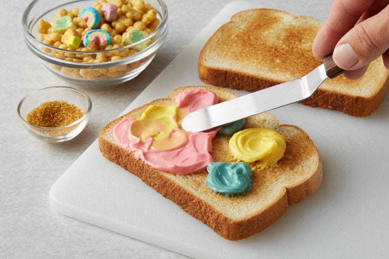 Untando Lucky Charms™ rainbow-colored spread en una tostada espolvoreada con cereal Lucky Charms™ Honey Clovers.
