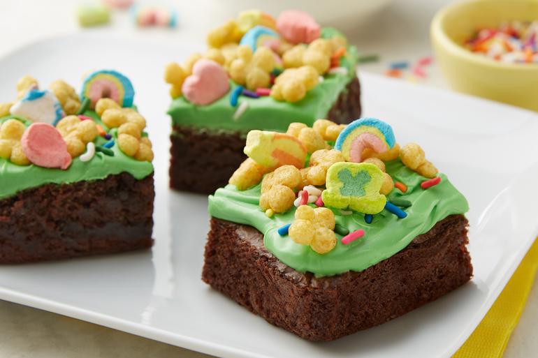 Tres trozos de Brownies glaseados de Lucky Charms en un plato blanco con cereal Lucky Charms™ en un plato blanco.