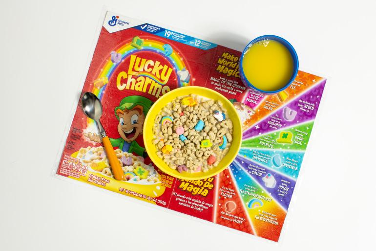Un pastel de capas arco iris del duende Lucky Charms con una quinta parte de la torta quitada y cereales Lucky Charm derramandose de su interior. Detrás de la tarta hay platos y un tenedor.