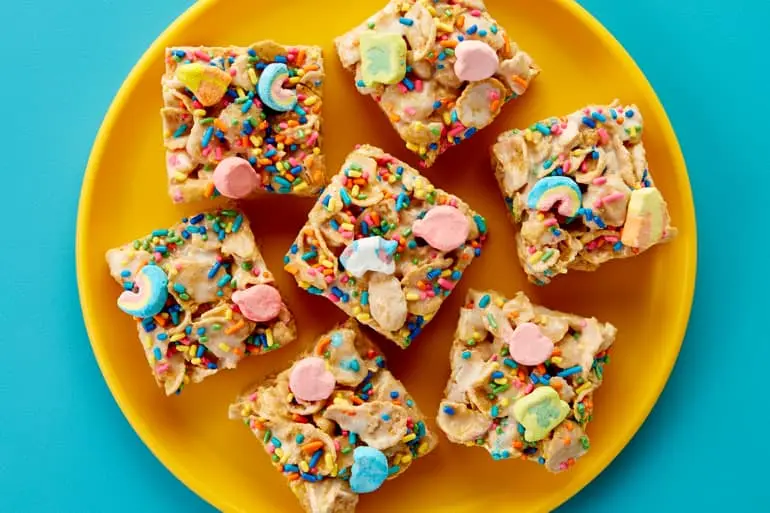 Overhead shot of 7 No-Bake Lucky Charms™ Frosted Flakes Cereal Bars on a yellow plate with blue background.