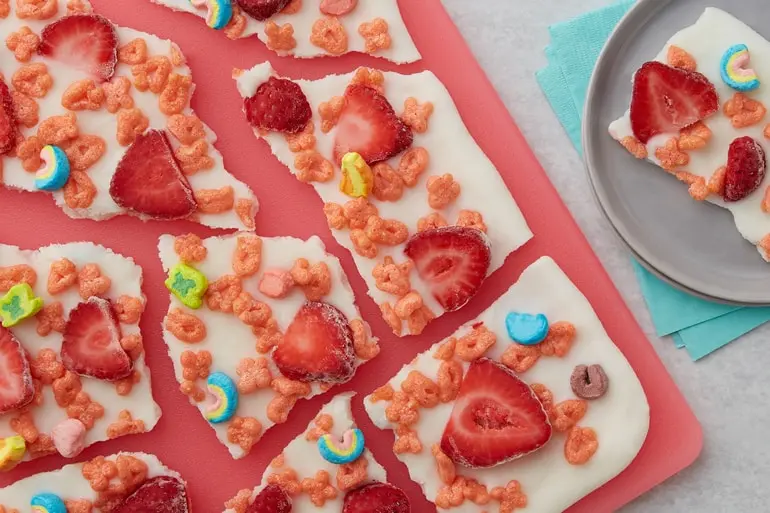 Broken up Frozen Yogurt Bark topped with Fruity Lucky Charms™ cereal and sliced strawberries on a cutting board.