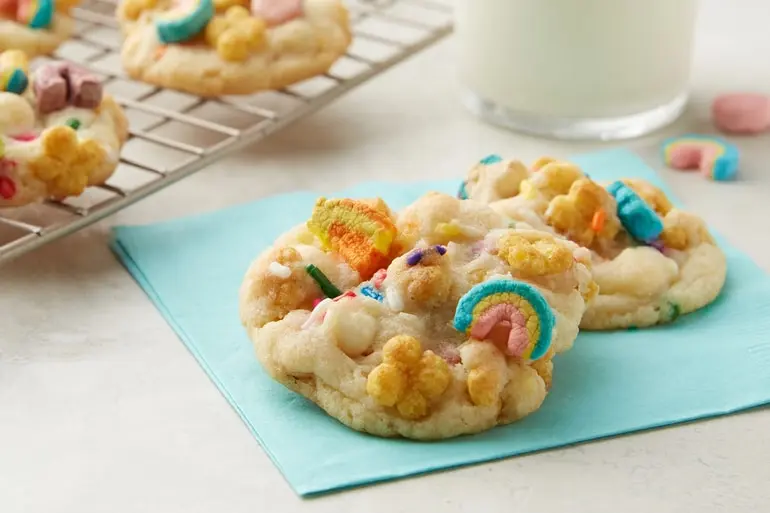 Two cereal cookies sprinkled with Lucky Charms™ Honey Clovers cereal presented on a blue napkin.