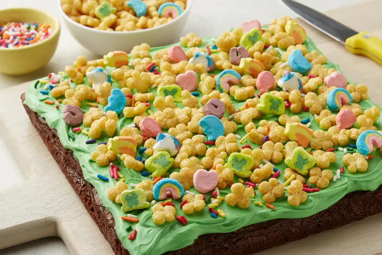 Unsliced Frosted Brownie recipe topped with Lucky Charms™ Honey Clover cereal served on a cutting board.