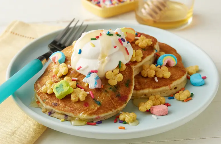 Lucky Charms™ Honey Clovers Cereal Pancake recipe with whipped cream, honey and sprinkles on a white plate.