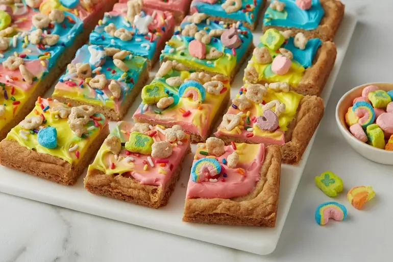 Sliced Rainbow Frosted Cookie Bars topped with Lucky Charms™ cereal on a cutting board.