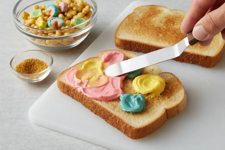 A hand spreading Lucky Charms™ rainbow-colored cream cheese spread on toast.