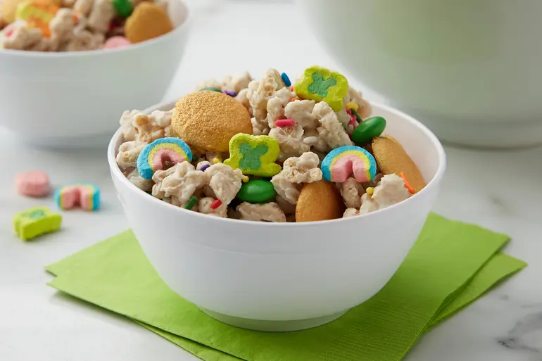Lucky Charms™ Leprechaun Gold Snack Mix topped with vanilla wafer cookies in a white bowl on a green napkin.