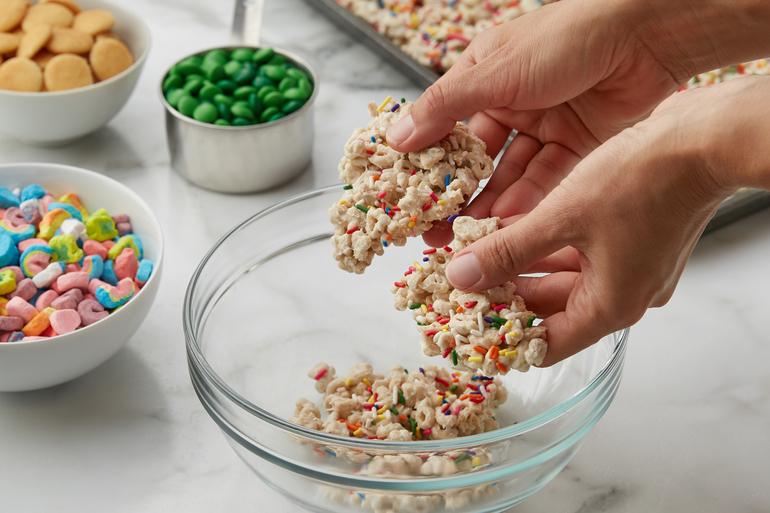 Two hands preparing Lucky Charms™ Leprechaun Gold Snack Mix in a glass mixing bowl.