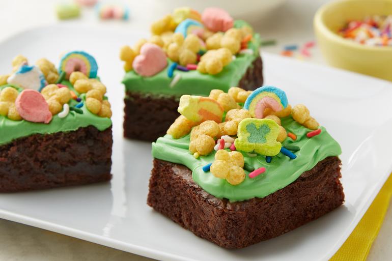 Three squares of green-frosted brownies topped with Lucky Charms™ Honey Clover cereal on a white plate.