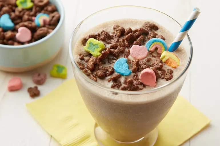 Lucky Charms™ Cereal topping a chocolate shake with straw in a tall glass beside a bowl of Chocolate Lucky Charms™.