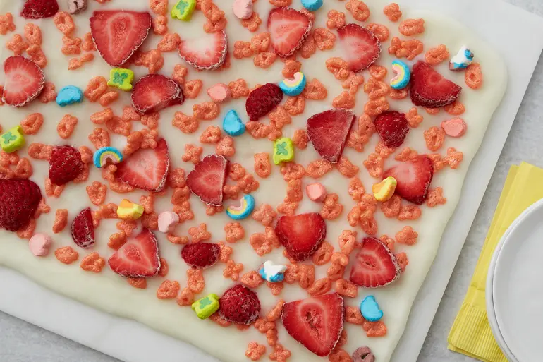 Unsliced Frozen Yogurt Bark topped with Fruity Lucky Charms™ cereal and sliced strawberries on a cutting board.