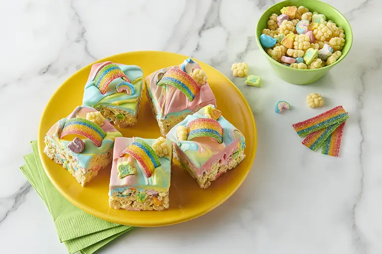 Five squares of Lucky Charms™ Frosted Rainbow Cereal Bars served on a round yellow plate on a marble counter.