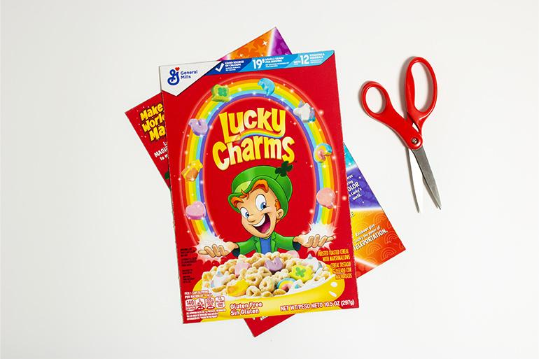 Lucky Charms™ Cereal Box Placemat Step 1 showing a cereal box cut into 2 parts and a scissors.