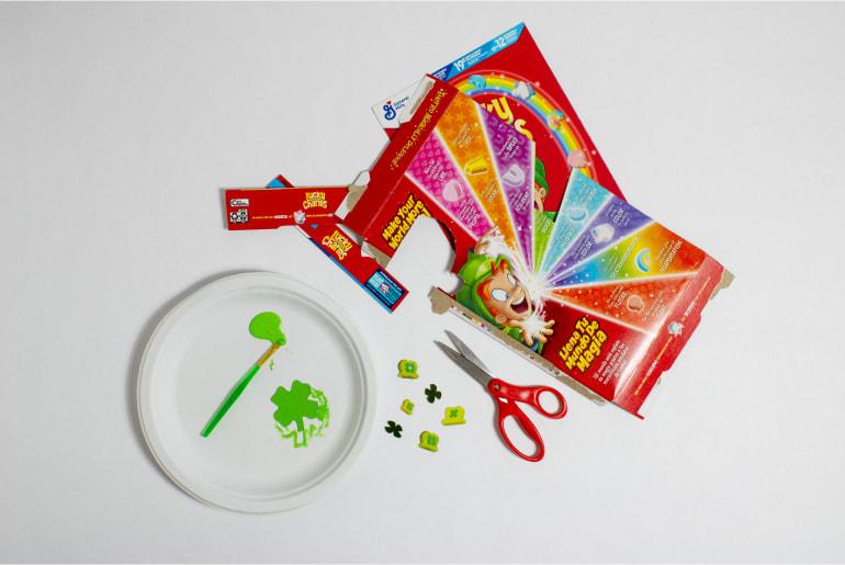 A Lucky Charms cereal box cut up, with scissors and a plate with paint and a paintbrush on top of it.