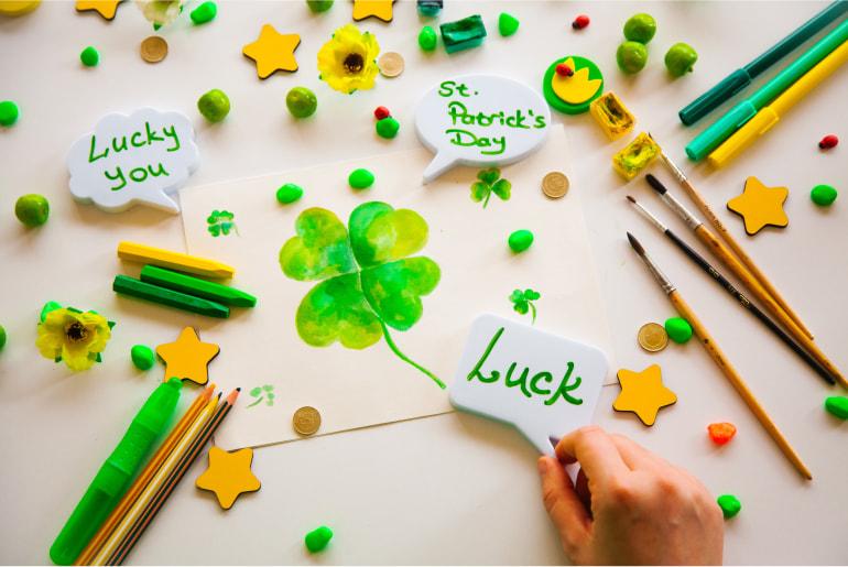 A painting of a 4 leaf clover with stars, pens, pencils, brushes and three labelled signs around the painting relating to luck.