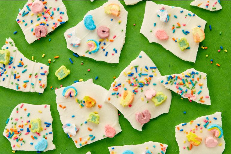 A table of frozen yogurt bark broken up into irregular pieces, each piece decorated with Lucky Charms, rainbow sweets and sprinkles.