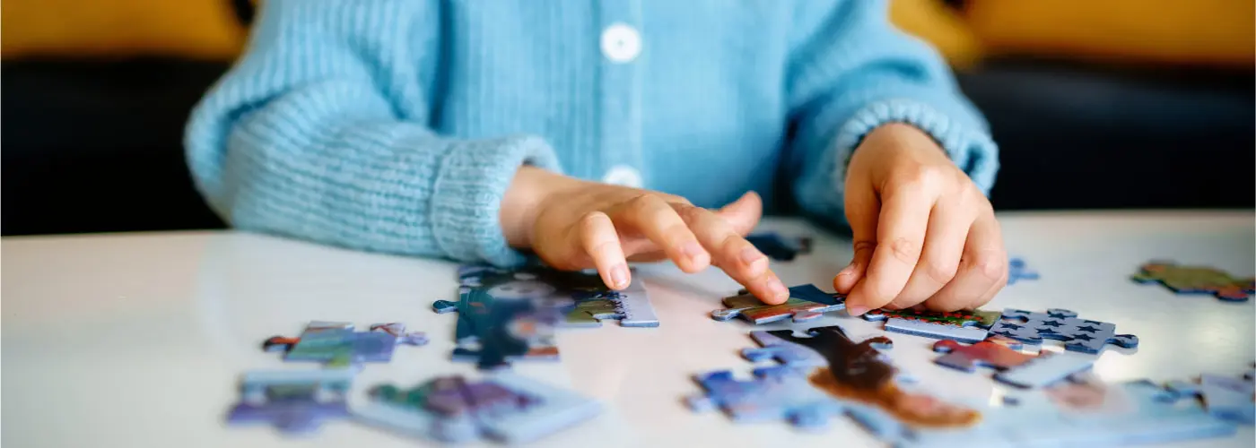 A child holding a jigsaw puzzle piece with many more puzzle pieces put together and scattered round a table.
