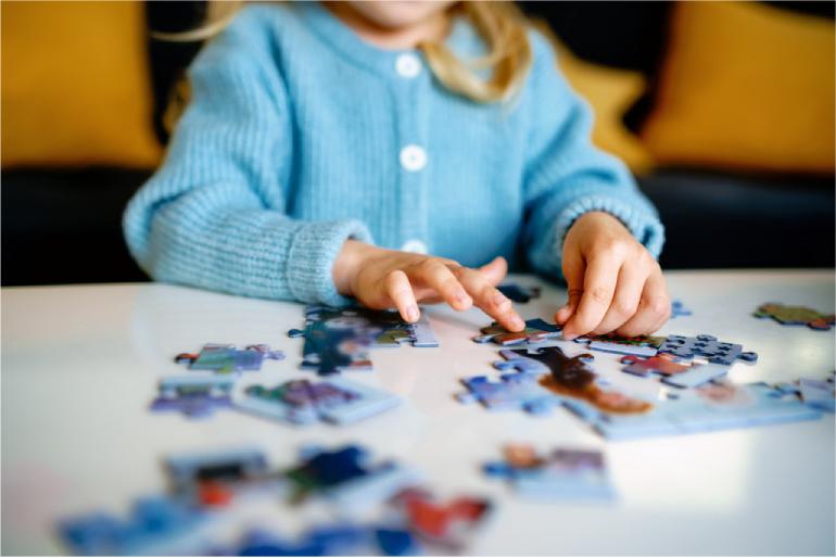 A child holding a jigsaw puzzle piece with many more puzzle pieces put together and scattered round a table.
