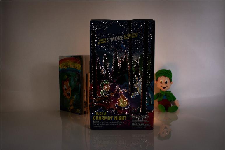 A Lucky Charms cereal box decorated with LED lights with "The Magic Inside" book behind it and a Lucky plush toy beside it.