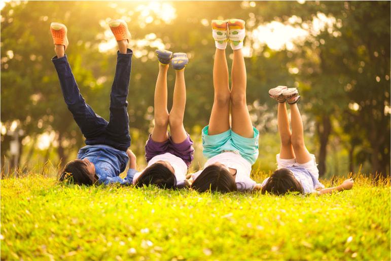 Four kids lying down in a row on some grass pointed away from the camera with their legs up in the air on a sunny day.