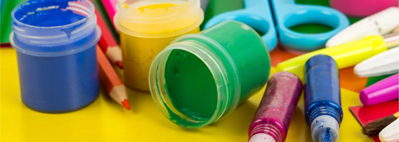 Colorful craft supplies lying on a table, including glitter pens, pots of paint, colored paper and pencils.