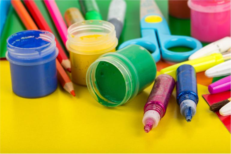 Colorful craft supplies lying on a table, including glitter pens, pots of paint, colored paper and pencils.