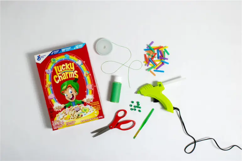 A Lucky Charms cereal box on a table with craft supplies beside it, including scissors, thread, a glue gun and beads.