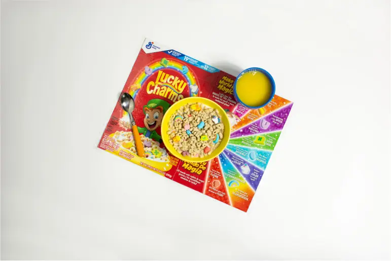 A Lucky Charms cereal box opened to be used as a placemat with a bowl of Lucky Charms cereal and a glass of orange juice on it.