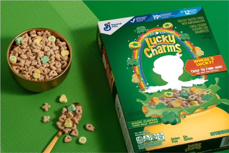 A green box of Lucky Charms cereal lying on a green table beside a gold bowl of Lucky Charms cereal and spoon.