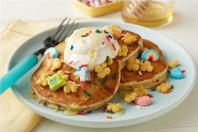 A blue plate of homemade pancakes topped with whipped cream, Lucky Charms pieces, sprinkles and honey, with a blue fork beside it.